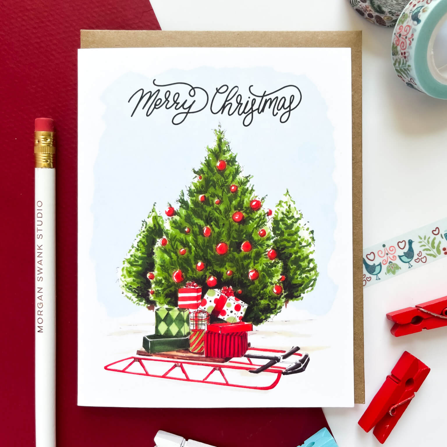 Holiday card showing a decorated Christmas tree with red ornaments and wrapped presents on a red sled, with the text 'Merry Christmas.'