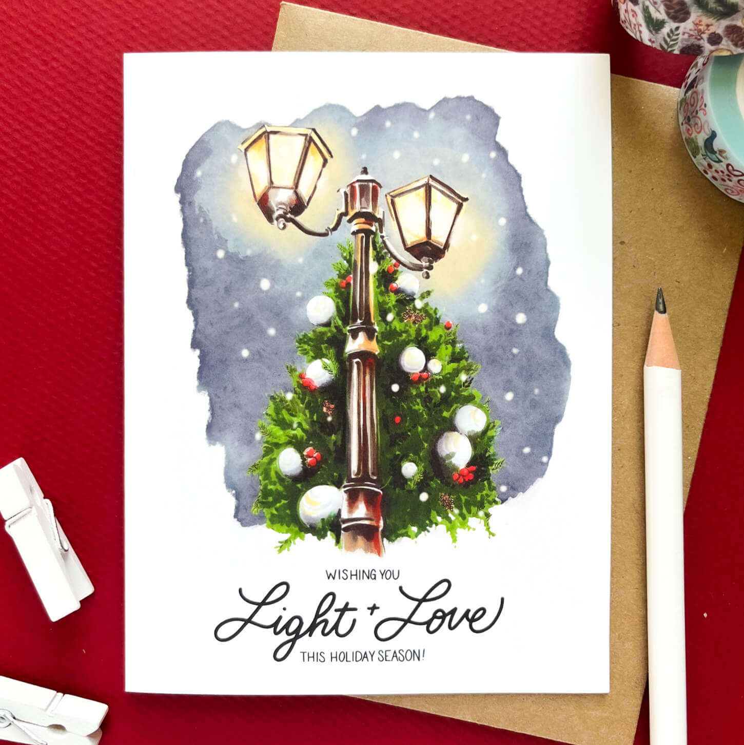 Christmas card featuring a glowing vintage streetlamp surrounded by a decorated evergreen tree, with the text 'Wishing you light + love this holiday season.'