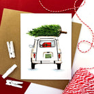 Holiday card showing a white retro car from the back, with a Christmas tree on the roof and gifts in the trunk.